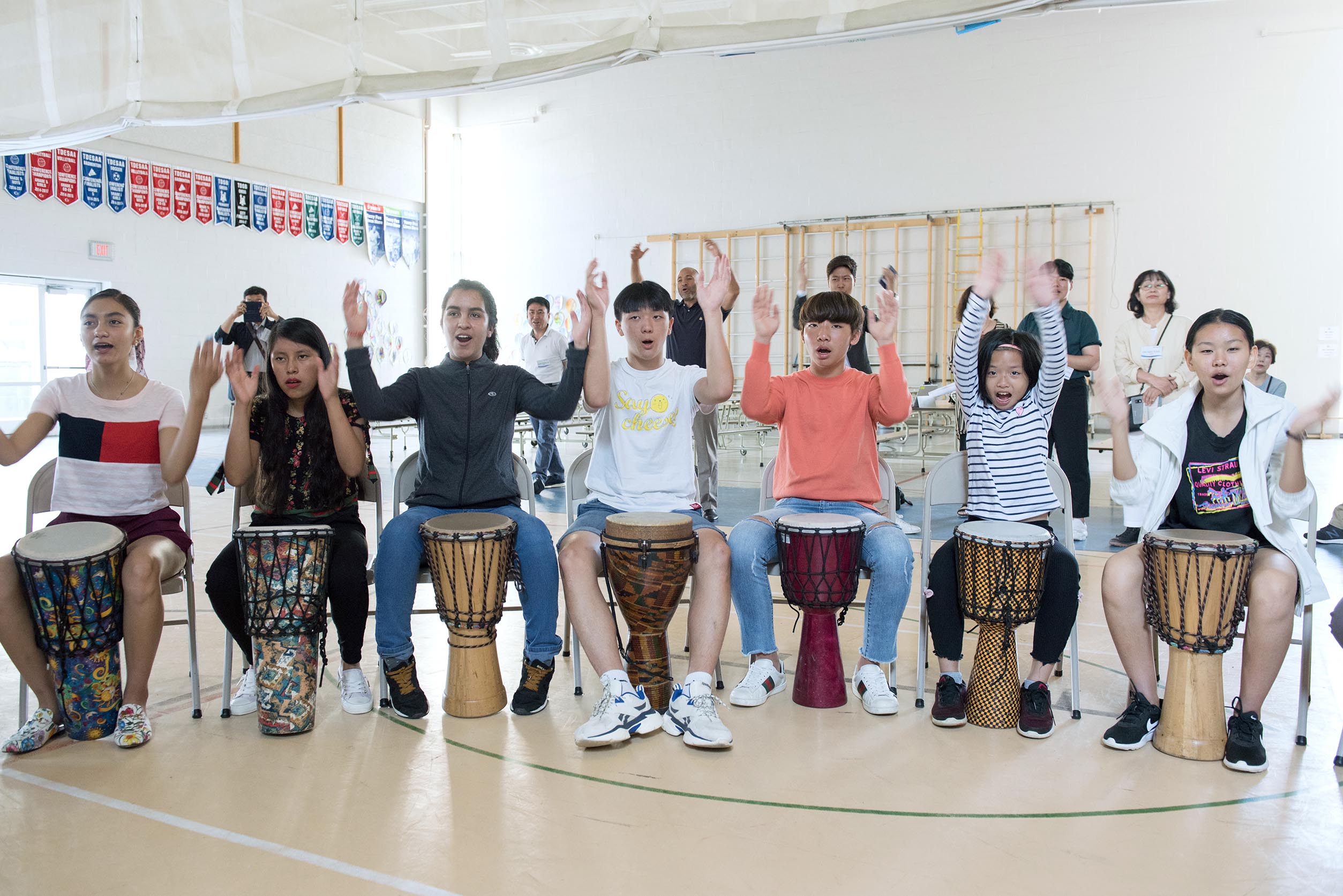 Five students sitting side by side with their hands in the air playing drums Open Gallery